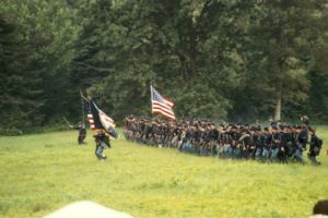 A group of costumed reenactors marching in a line with flag-bearers carrying American flags.