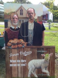 A life-size copy of the "American Gothic" image of a farm couple, with cut-outs for people to pose and two men looking through the cut-outs.
