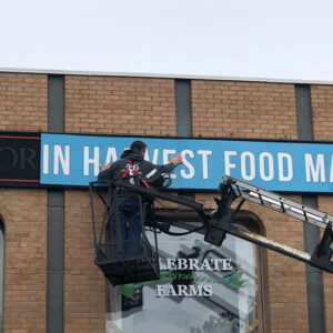 Man in a lift installing a partially readable sign, "-in Harvest Food M"